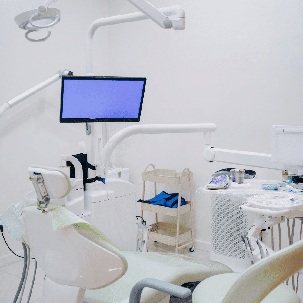 A modern dental clinic room with a dental chair, overhead light, monitor, instrument tray, and medical cart in a clean, white setting.