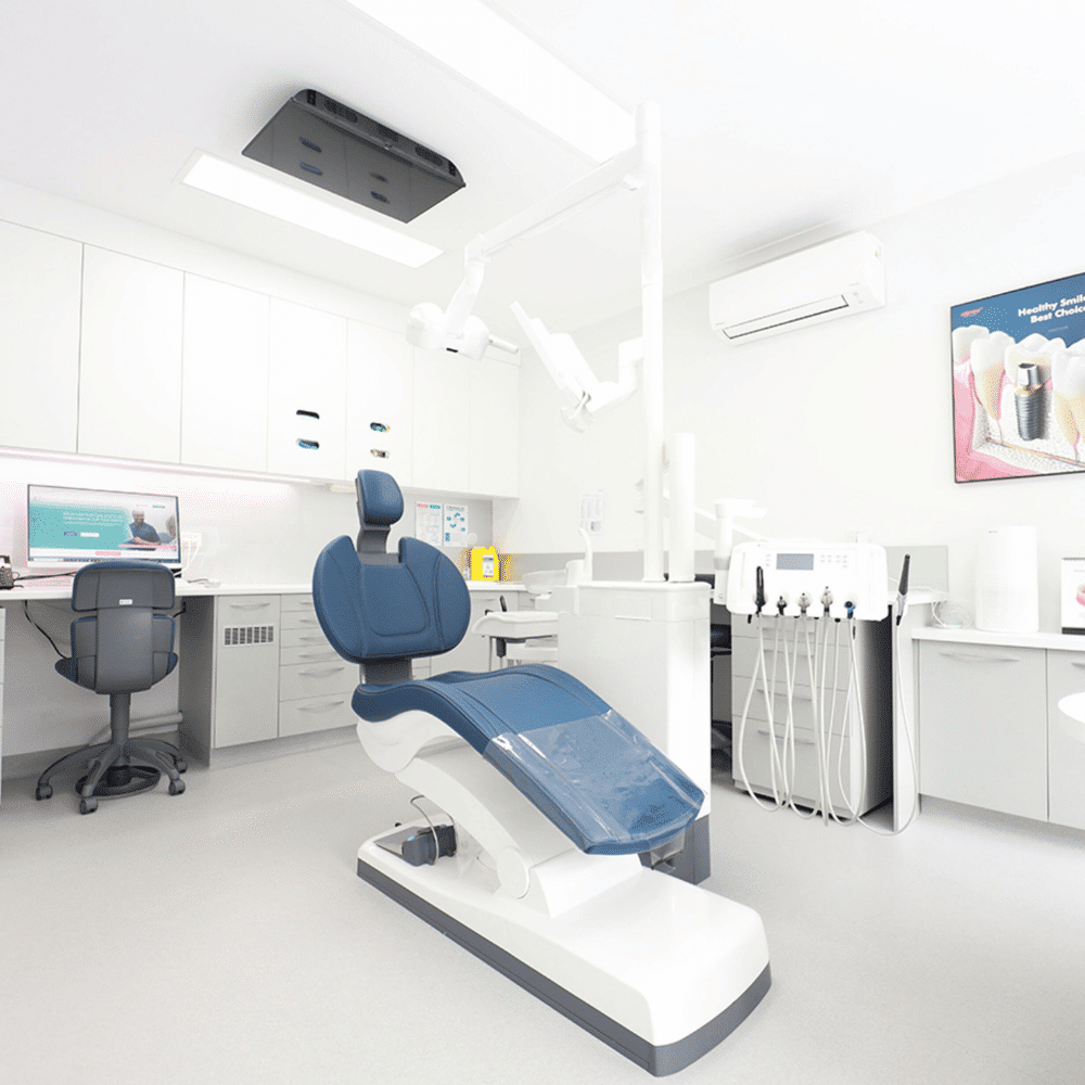 A modern, clean dental office with a blue dental chair in the center, dental equipment nearby, a computer on a desk, white cabinets, and bright lighting.