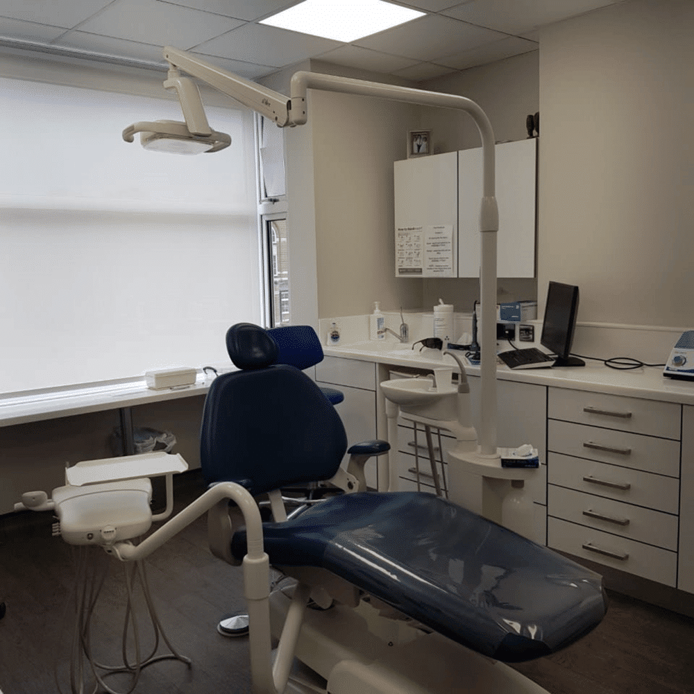 A modern dental office with a blue dental chair, overhead exam light, white cabinets, a computer on the counter, and dental equipment organized around the room. The space is clean and well-lit with natural light.