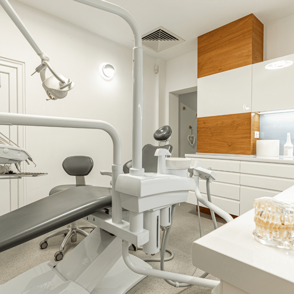A modern dental office with a reclining dental chair, overhead light, drawers, cabinets, and dental tools. The space is clean, bright, and organized with white and wood finishes.