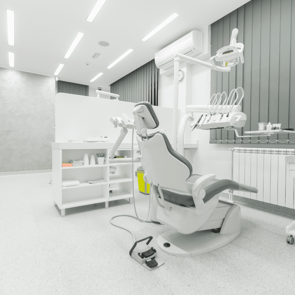 A modern, empty dental office with a gray and white dentist chair, overhead light, and dental instruments next to a counter with supplies, all in a brightly lit, clean environment.