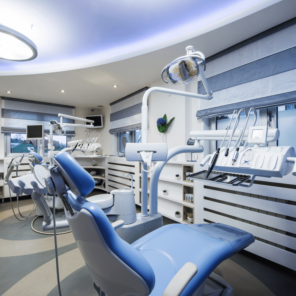A modern dental clinic with blue dental chairs, overhead lights, and various dental instruments organized on white shelves, featuring large windows and a clean, bright interior.